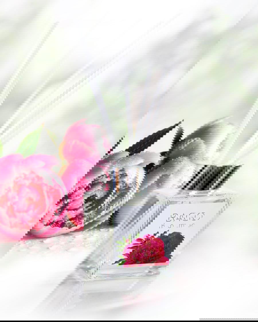 Стеклянный диффузор с палочками и этикеткой с пионом на светлом фоне, рядом живые цветы — идея ароматического подарка для дома