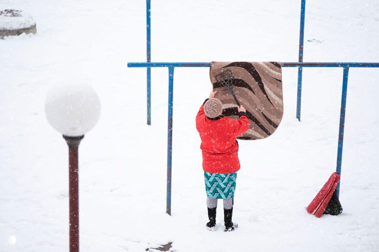 Человек подвесил ковёр на перила и выбивает его, вокруг идёт снегопад; видно рабочий приём с подвешиванием и встряхиванием ворса.