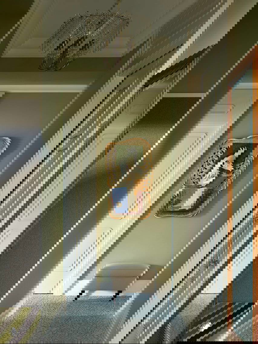 Классическая зелёная прихожая с высоким потолком: хрустальная люстра, декоративная настенная скульптура и небольшой пуф у стены.