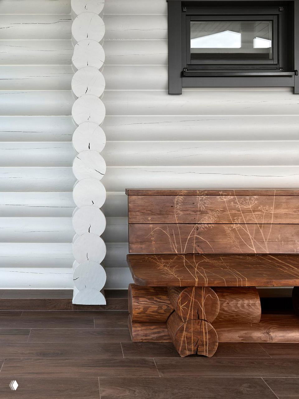 Деталь интерьера — деревянная скамья с декоративным резным рисунком на фоне белёной бревенчатой стены внутри дома.