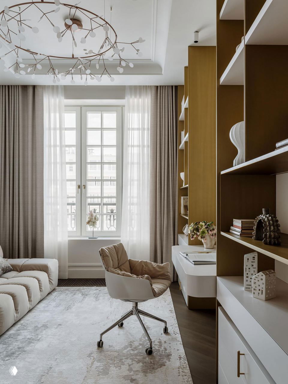 Рабочая зона/кабинет у окна с белым письменным столом, мягким креслом на колёсах, встроенными латунными стеллажами и коллекционными предметами декора на полках.