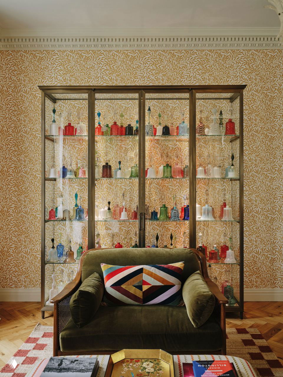 Витрина с коллекцией разноцветных стеклянных колокольчиков на полках на фоне растительных жёлтых обоев и бархатное кресло на переднем плане.