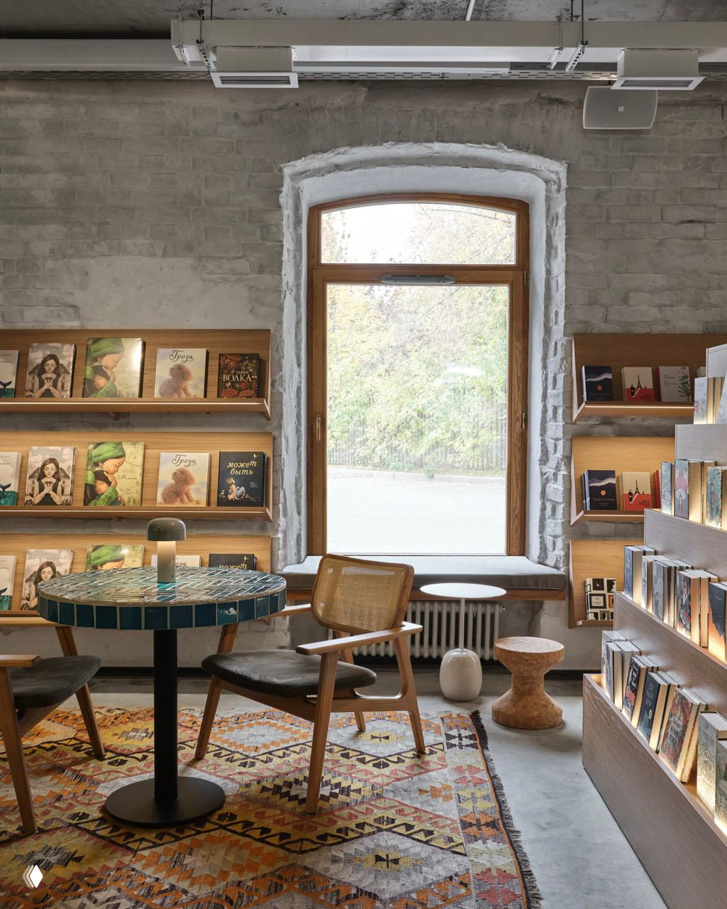 Небольшая уютная читальня у окна: кирпичная отделка, окно с видом на зелёную зону, полки с изданиями и кресло для спокойного чтения в дневном свете.