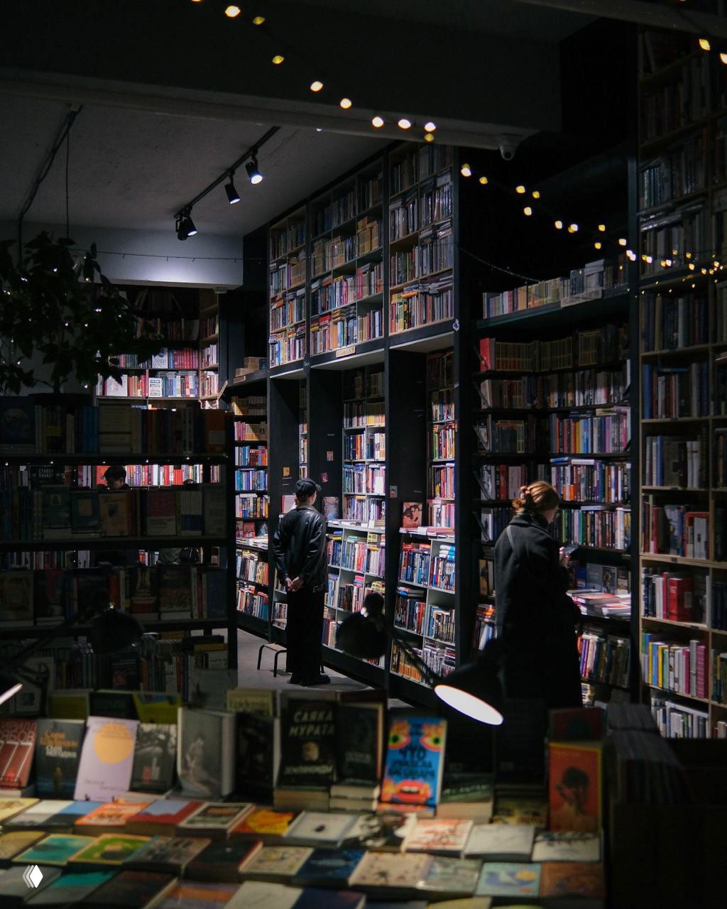 Тёмный книжный зал с высокими стеллажами, мягким локальным освещением и посетителями, выбирающими книги — атмосферная сцена вечернего книжного пространства.