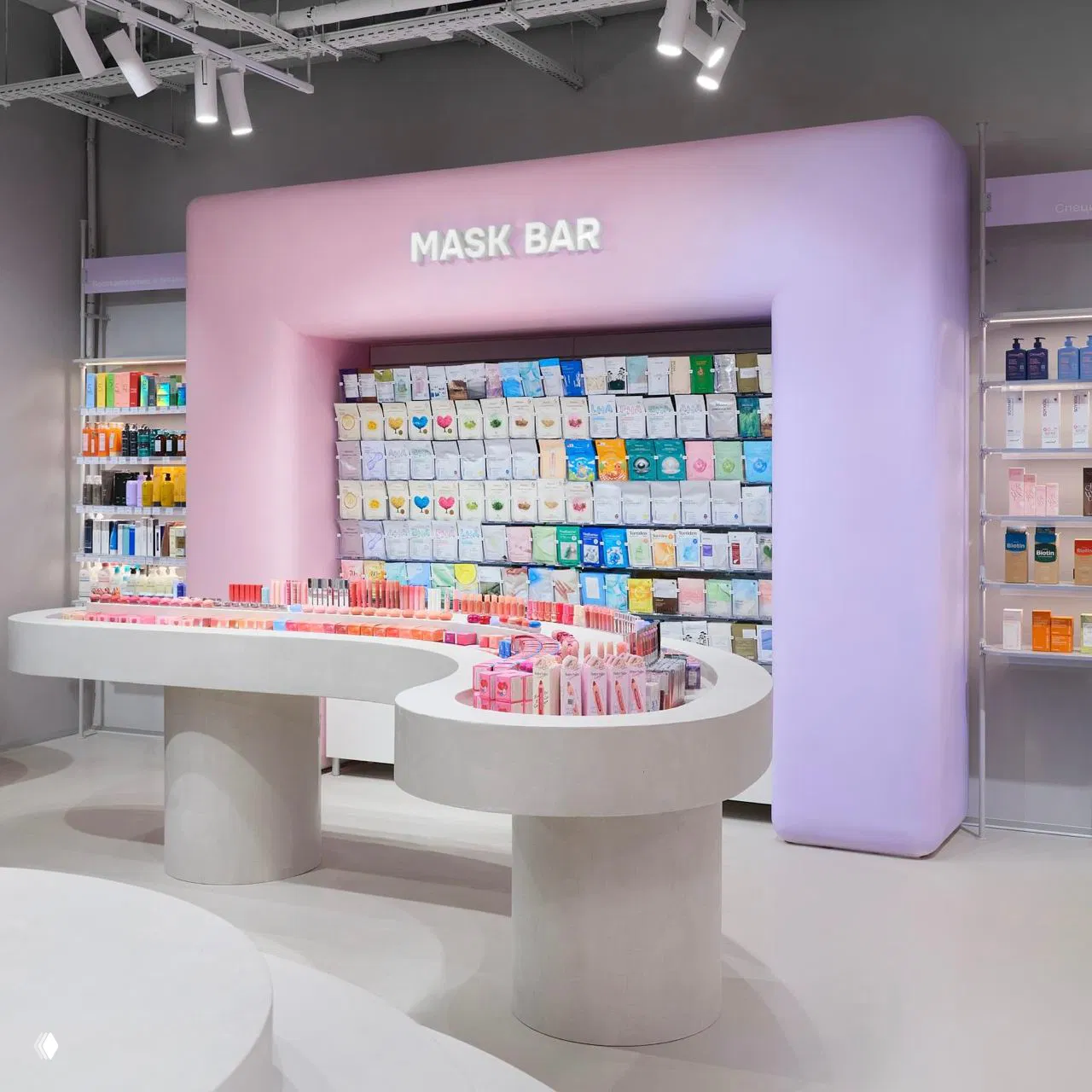 Крупный план изогнутого стола и стенда «MASK BAR» в лилово-розовой зоне магазина Hollyshop, расставленные средства по уходу, помады и миниатюры.