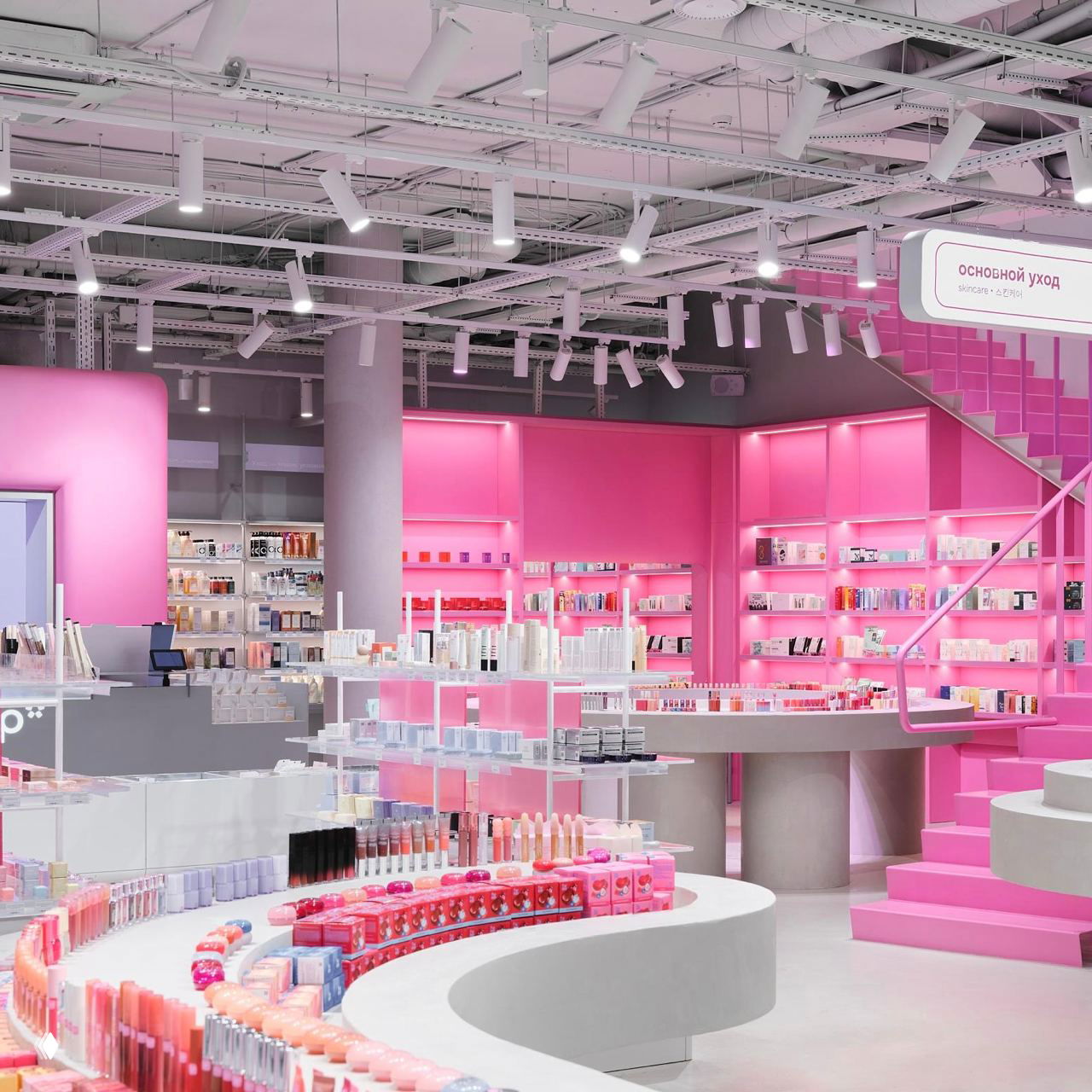 Широкий вид на двухэтажный уровень магазина: круговые столы, розовые стены, подсветка и полки с косметикой, лестница ведёт на камерный второй этаж.