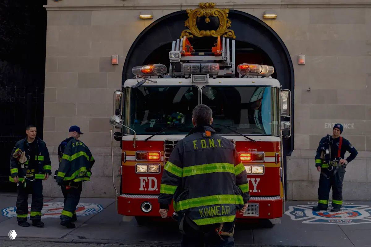 Пожарный грузовик FDNY выезжает из гаража депо, на переднем плане пожарный в защитной куртке с буквами FDNY на спине