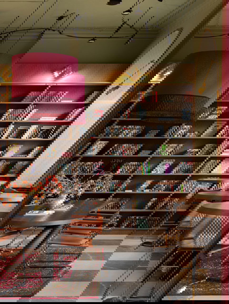 Читальная зона с деревянными стеллажами, обилием книг, длинным столом и красными акцентами: ковёр под столом, кресла и мягкое освещение создают домашний книжный салон.