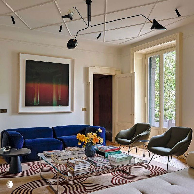 Гостиная с синим бархатным диваном, стеклянным журнальным столом и креслами у высоких декоративных окон, зелёный платан видно за окном.