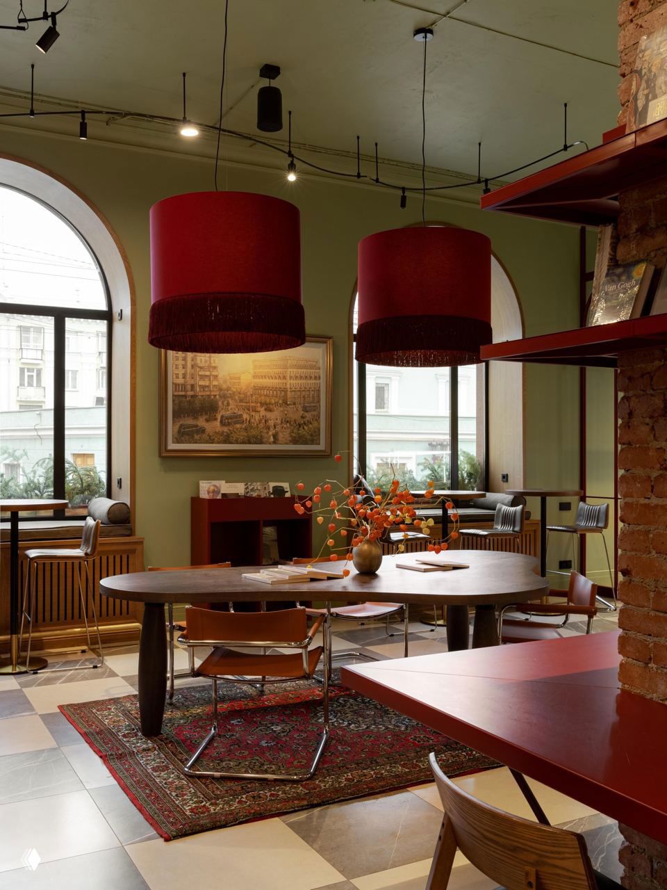 Большие арочные окна, два крупных красных подвесных светильника и столы у окна: вид на интерьер магазина, где классические элементы сочетаются с яркими современными акцентами.