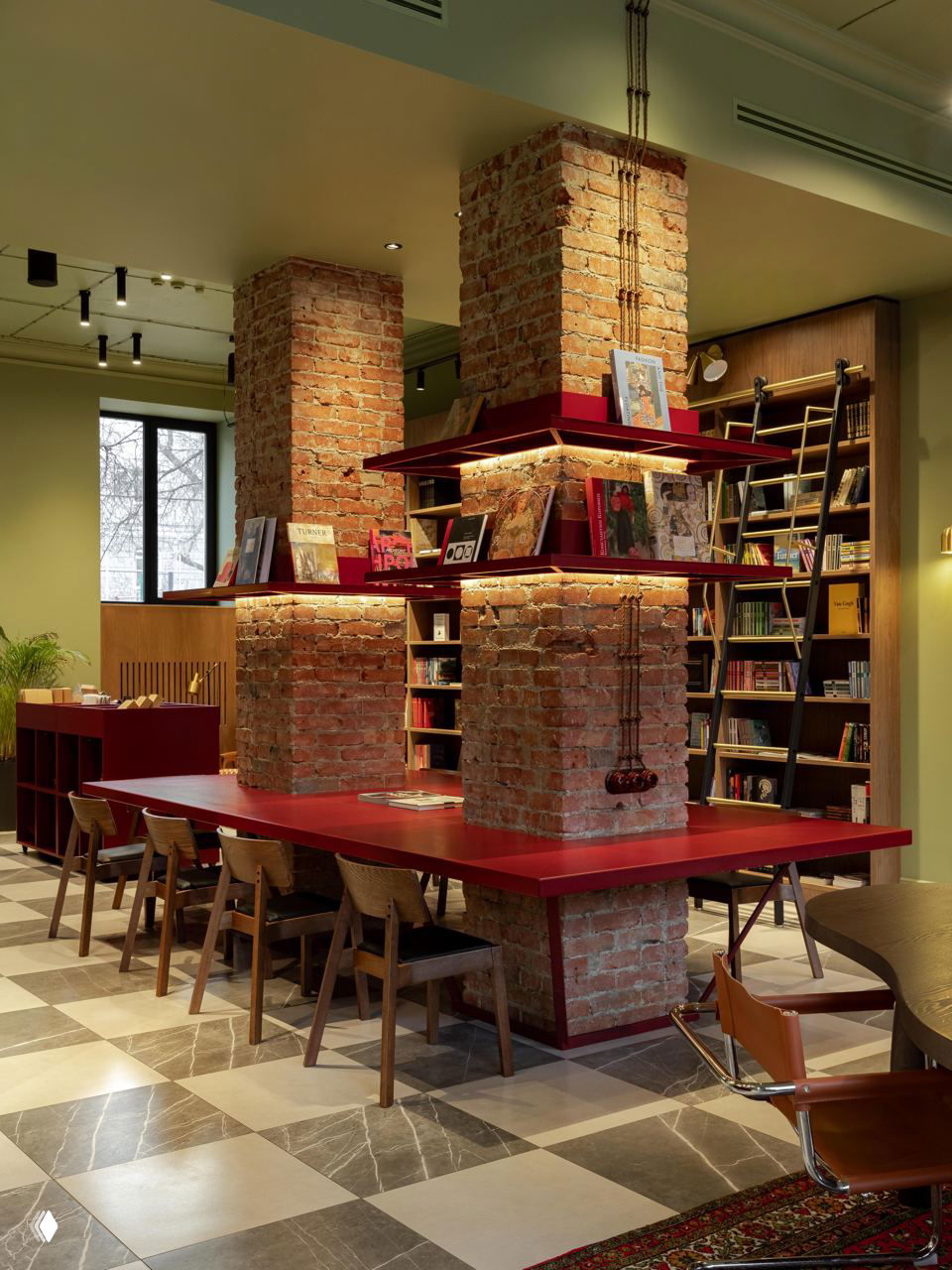Открытые кирпичные колонны, встроенные книжные полки и длинный красный стол вокруг колонн — индустриальные текстуры соседствуют с тёплой деревянной мебелью и декоративным освещением.