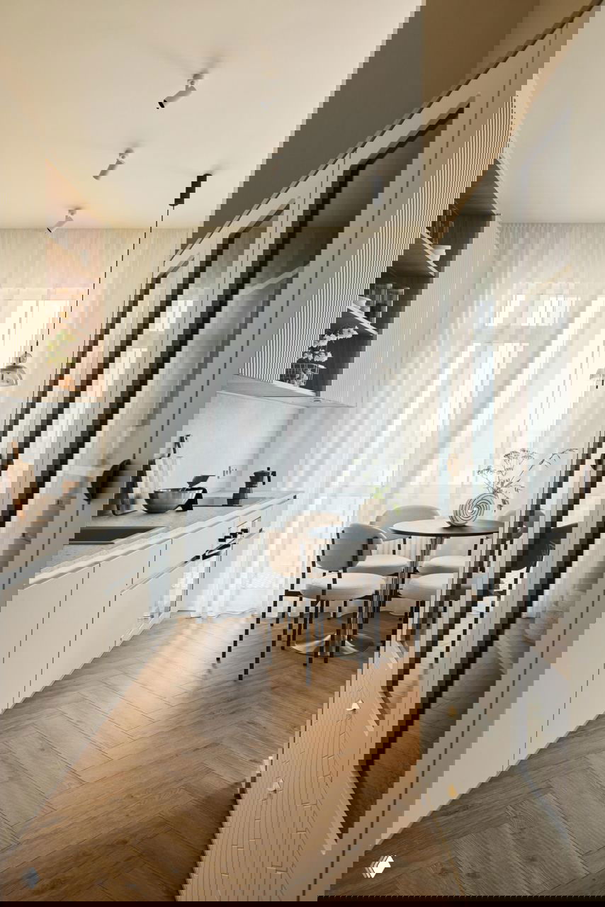 Вид на кухню и обеденную зону с высокими полупрозрачными шторами, небольшим круглым столом и стульями; панорамное окно добавляет свет и ощущение простора в мансардной квартире.