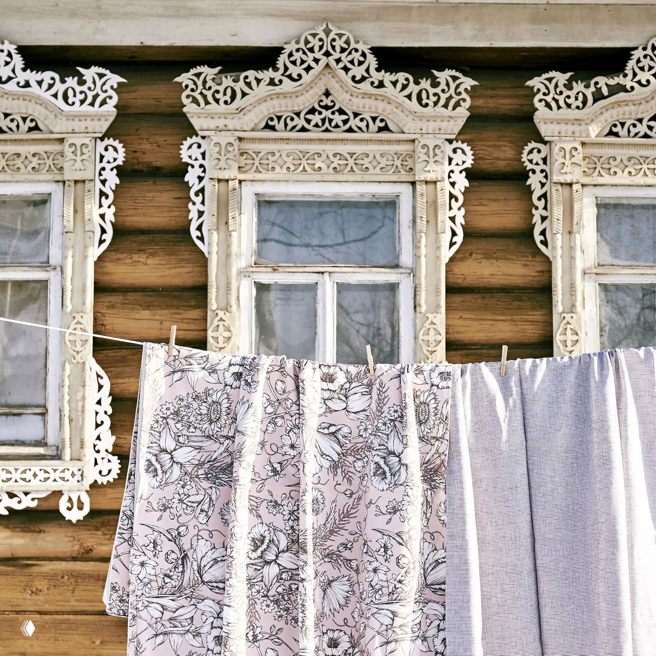 Сушащиеся на верёвке простыни и пододеяльники на фоне резной деревянной избы — традиционный способ проветривания и сушки текстиля.