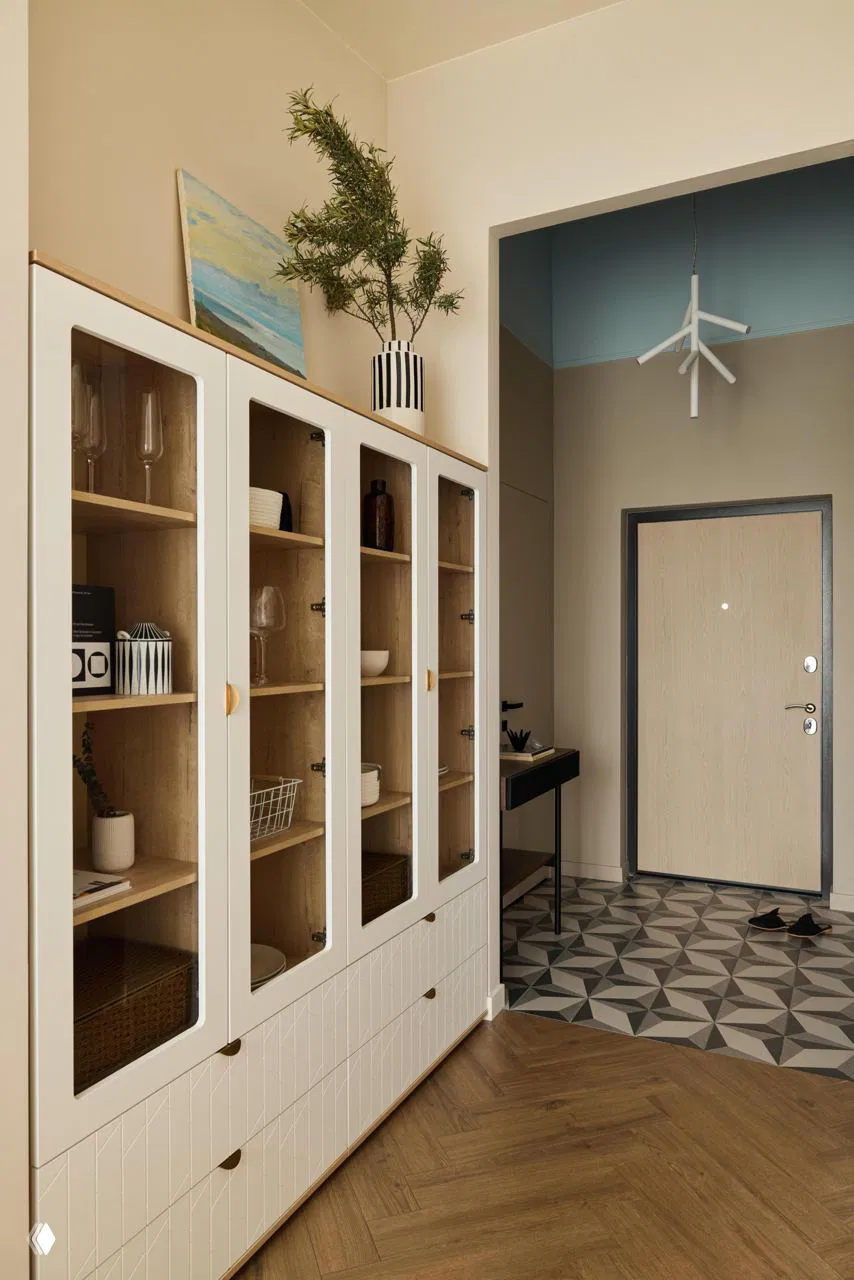 Прихожая с высокой витриной-буфетом в светлой отделке, декоративными предметами и геометрической плиткой у входа; тёплая нейтральная палитра и деревянные акценты формируют единый образ.