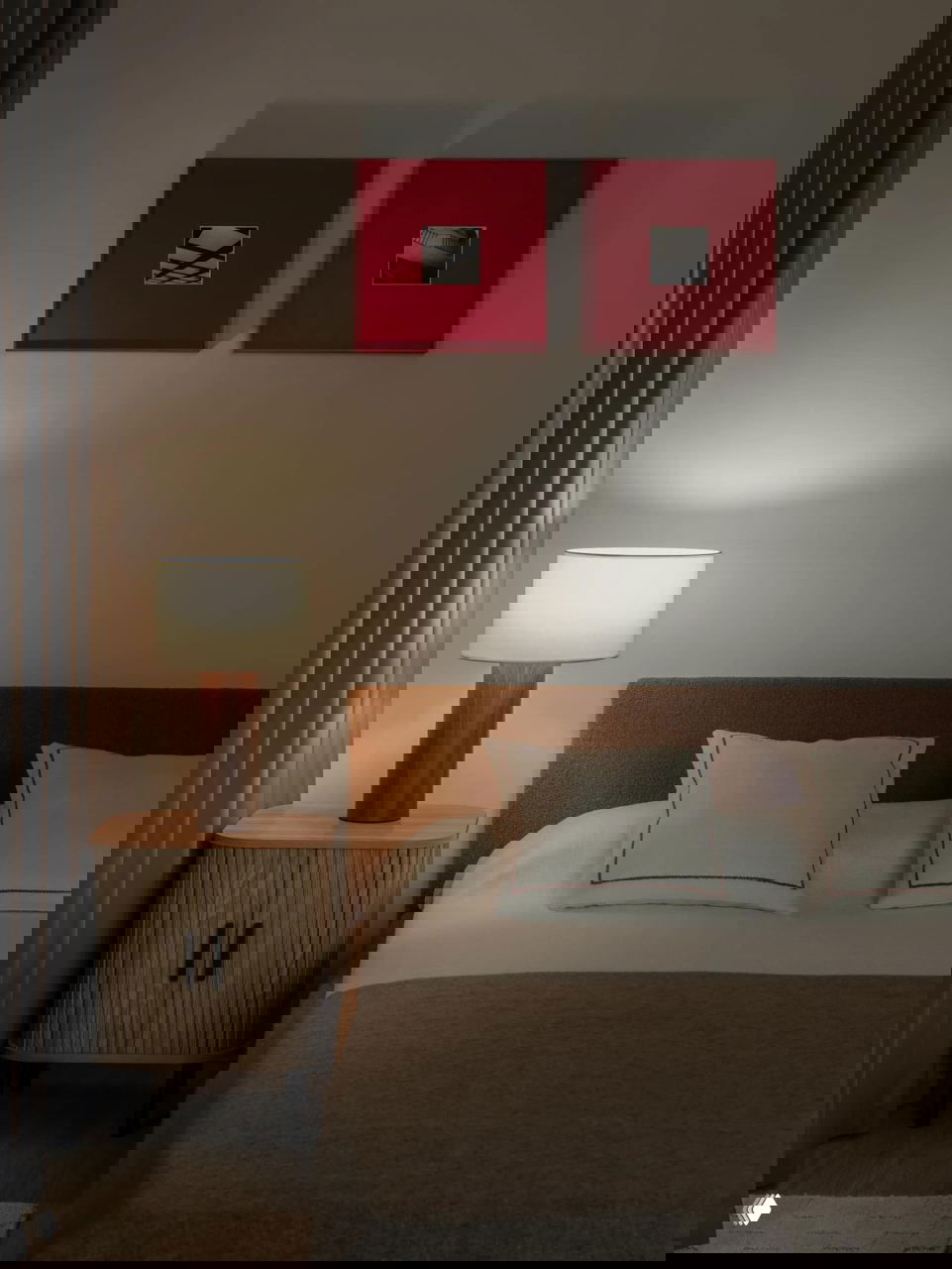 Уютная спальная часть с тёплым изголовьем, прикроватной тумбой и лампой; два ярких красных квадрата над кроватью выступают художественными акцентами в спокойной палитре.