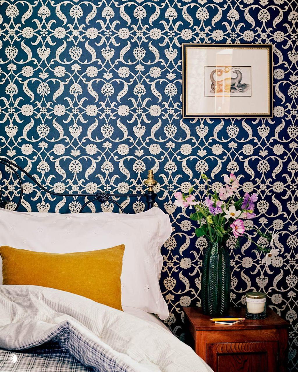 Деталь интерьера: глубокий синий обойный узор в викторианском стиле, кованая металлическая спинка кровати и прикроватный столик с вазой и цветами.