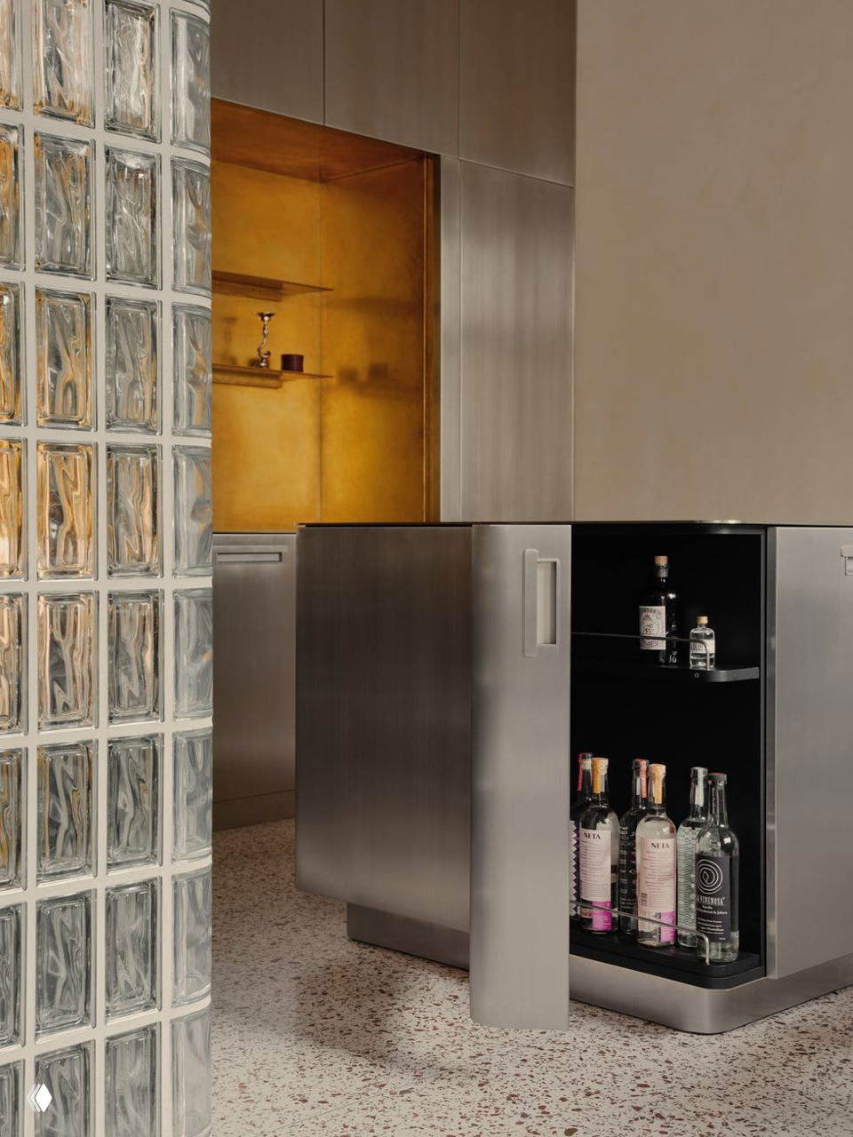 Кухонный блок из нержавеющей стали рядом со стеклоблоками; внутренняя латунная ниша и открытый шкаф с бутылками подчёркивают контраст материалов и фактур.
