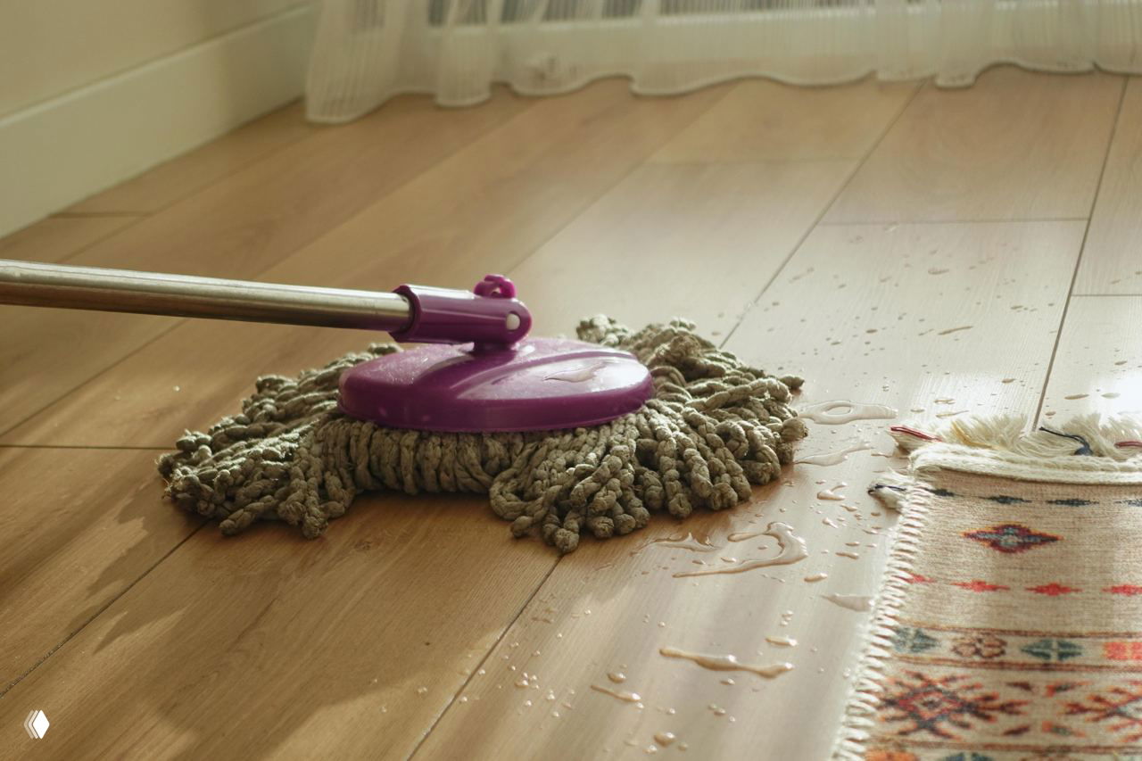 Моп на деревянном полу оставляет воду и следы рядом с ковриком — демонстрация риска избыточной влаги для паркета.