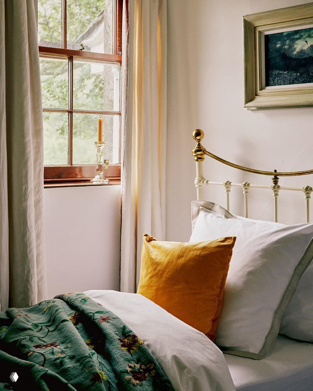 Спальня в старинном особняке: белая кровать с латунным изголовьем, жёлтая декоративная подушка и покрывало с цветочным рисунком рядом с окном и занавесками.
