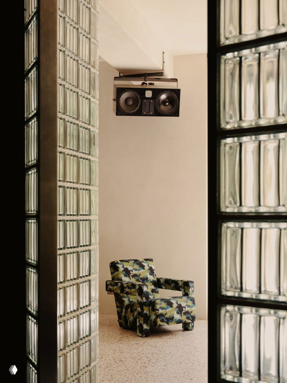 Проход между панелями из стеклоблоков к уютной зоне с креслом в пёстрой обивке и подвесной акустикой на потолке — клубная эстетика в жилом интерьере.