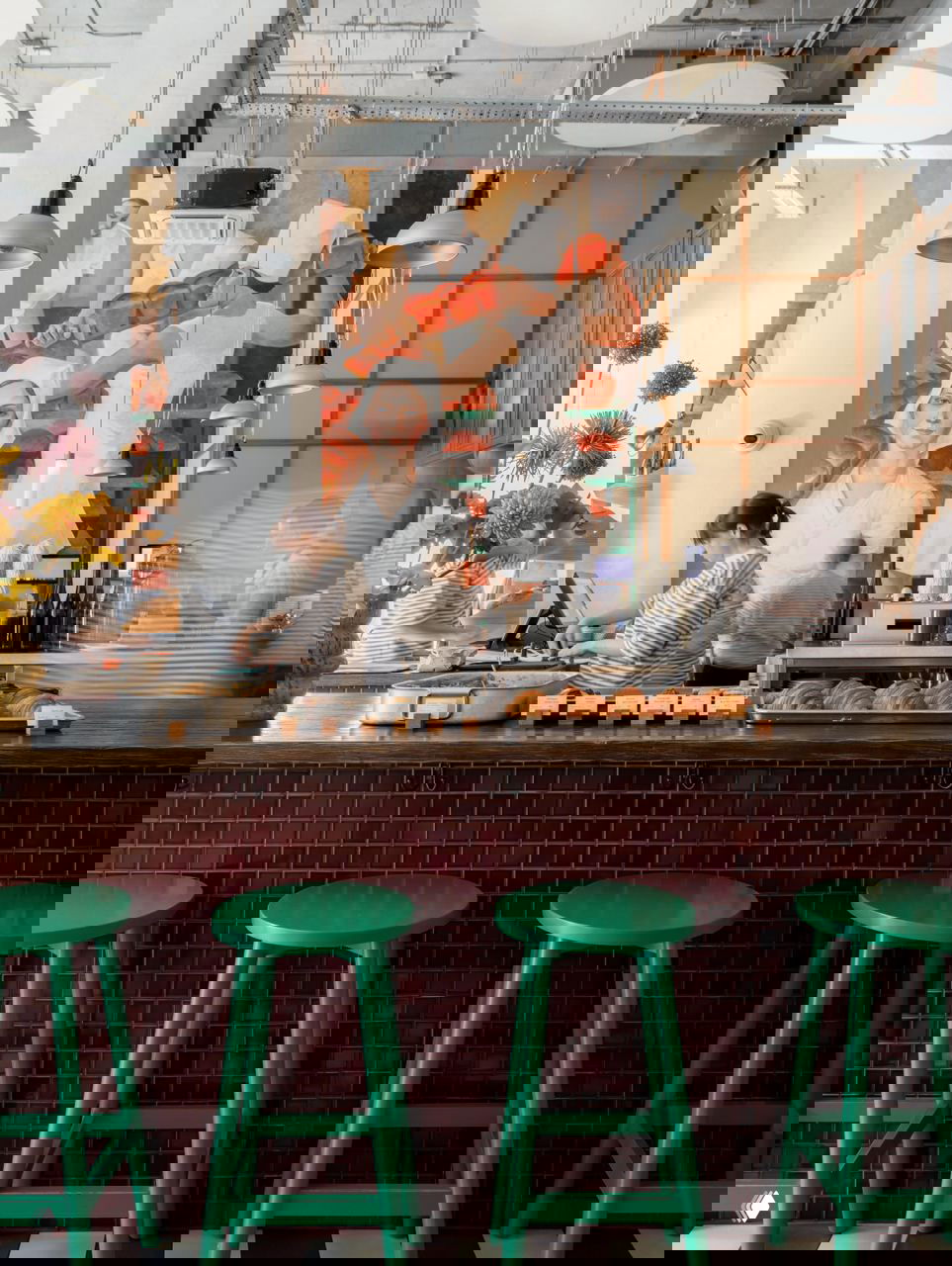 Стойка кафе с барменами и витриной круассанов, на заднем плане большая фреска в стиле соцреализма и ряд зелёных барных табуретов.