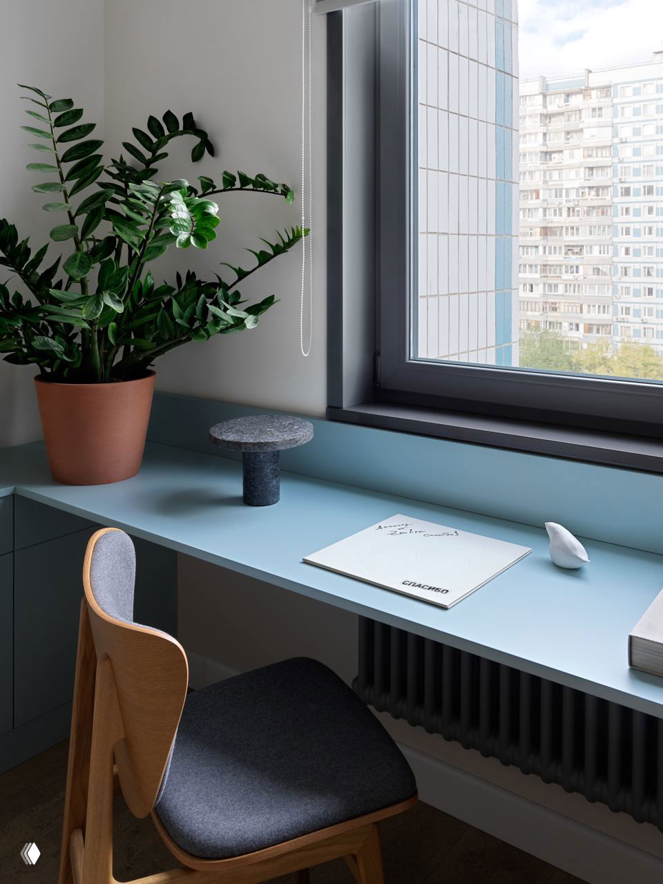 Рабочий уголок у окна: узкая голубая столешница, деревянный стул и горшечное растение — пример компактной и функциональной домашней рабочей зоны.