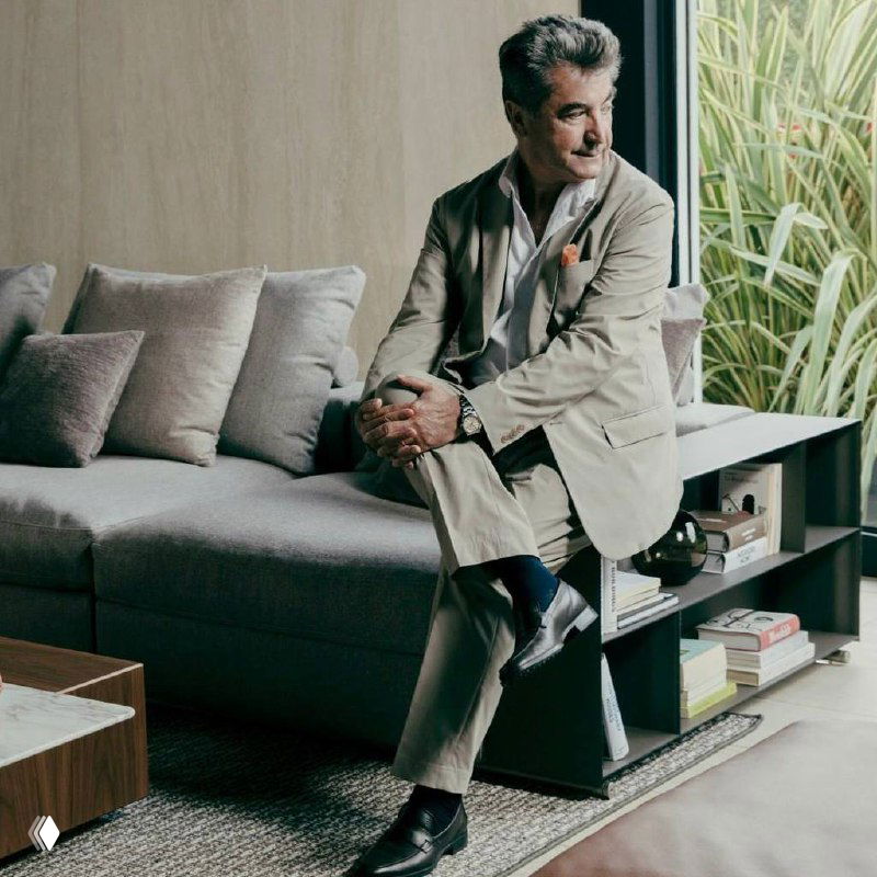 Портрет мужчины в костюме, сидящего на сером диване в современном интерьере: книги на полке, растения у окна, мягкие подушки