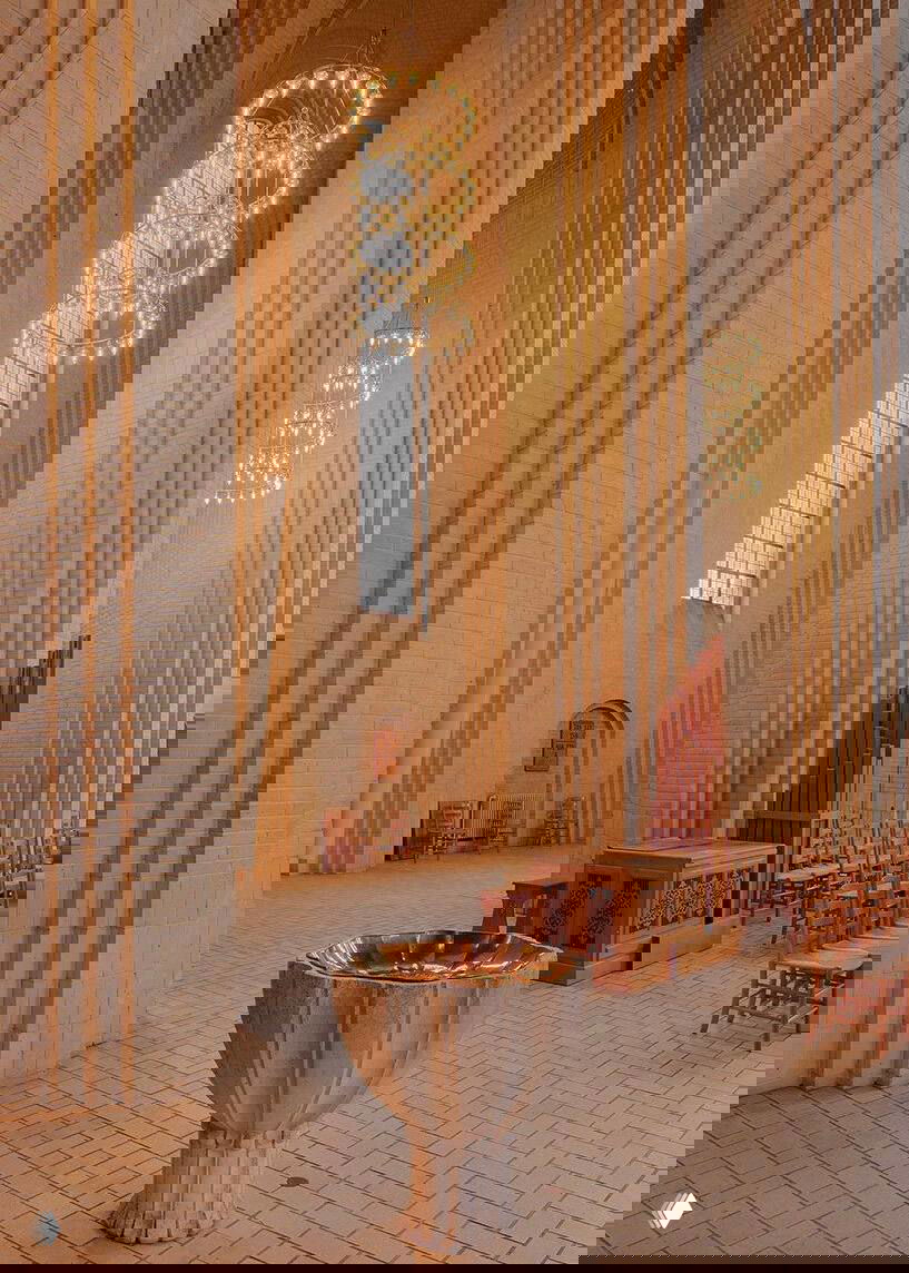 Крупный план литургического купели и окружающего интерьера церкви: кирпичное покрытие пола, деревянные детали и золотистое освещение люстр.