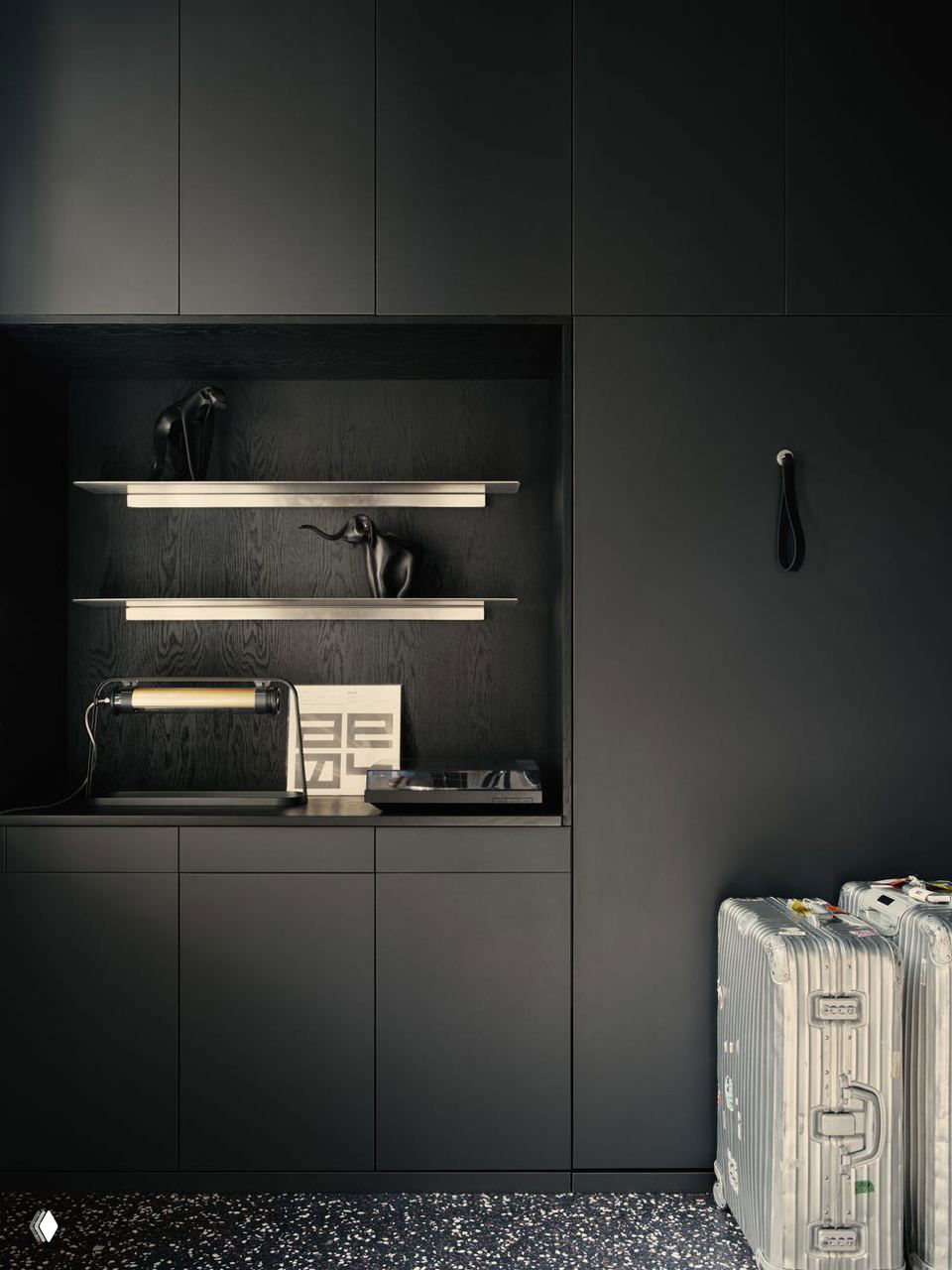 Темная встроенная мебель с открытыми полками, настольной лампой и металлическими дорожными чемоданами у стены; контраст матовых поверхностей и блеска металла на терраццо‑полу.