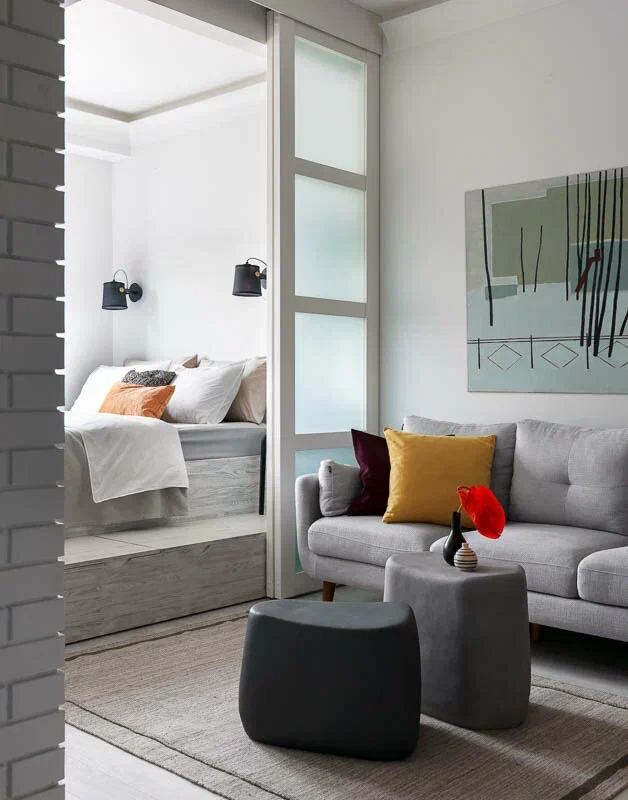 Небольшая гостиная с светло‑серым диваном, низкими пуфиками и открытым переходом в спальную зону — пример рационального зонирования маленькой квартиры.