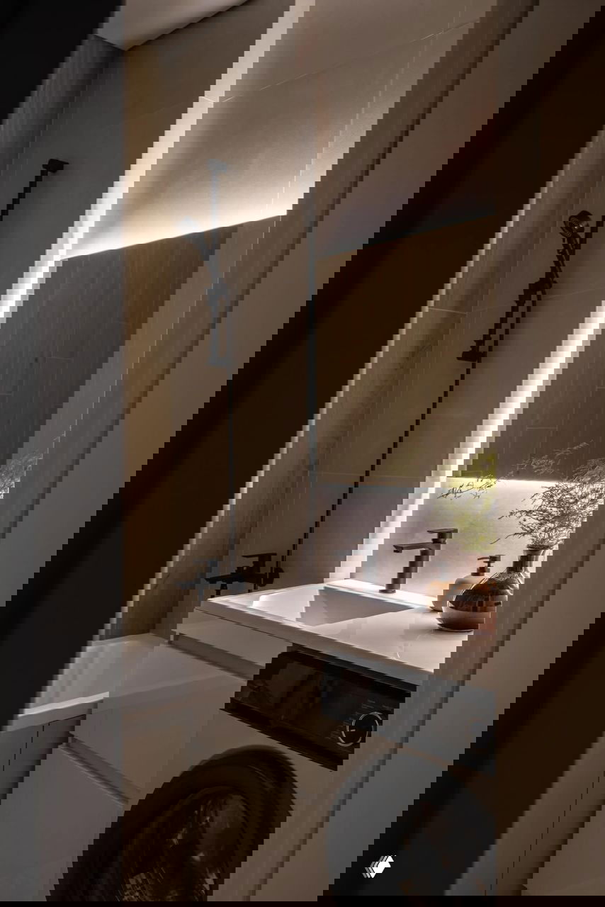 Небольшая ванная комната с душевой стойкой, подсвеченным зеркалом и встроенной стиральной машиной под раковиной, рациональное оформление для малогабаритной квартиры.