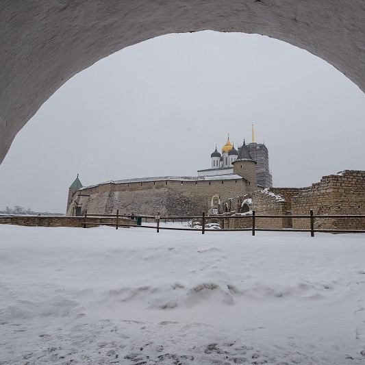 Зимний вид на Псковский кремль через арку: заснеженная земля, белые стены и силуэты Троицкого собора на фоне неба.