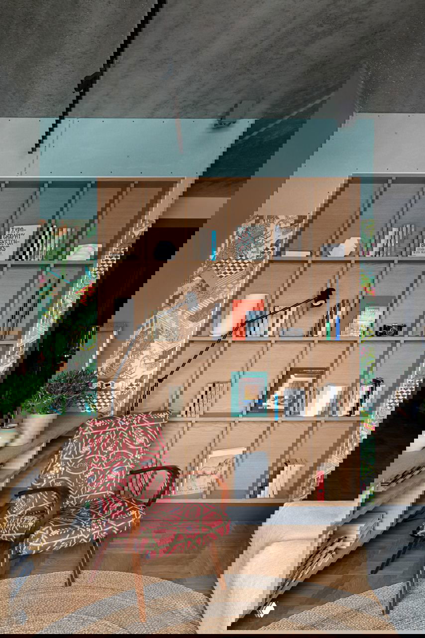 Узкая вертикальная стеллажная ниша с квадратными секциями, деревянный книжный шкаф в гостиной, кресло с орнаментом и декоративные обои по бокам.