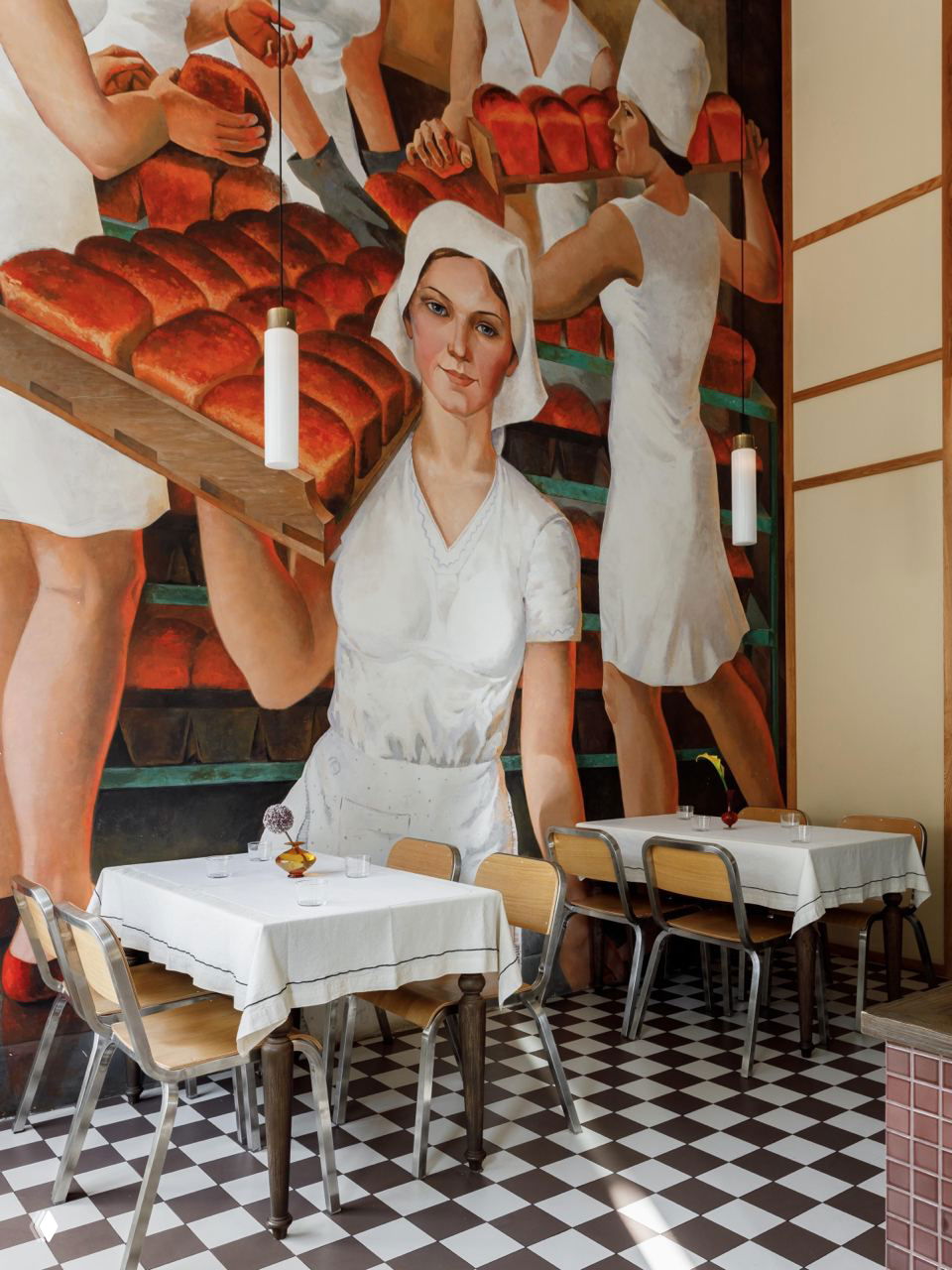 Крупный план фрески с изображением пекарей и тестом; возле стены расположены два накрытых белыми скатертями столика и стулья.