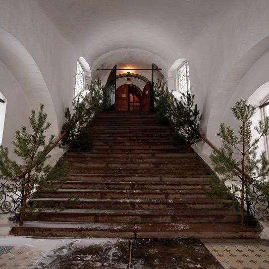 Интерьерная сцена внутри сооружений кремля: деревянная лестница, арочные окна и праздничная декорация из ёлок у стен.