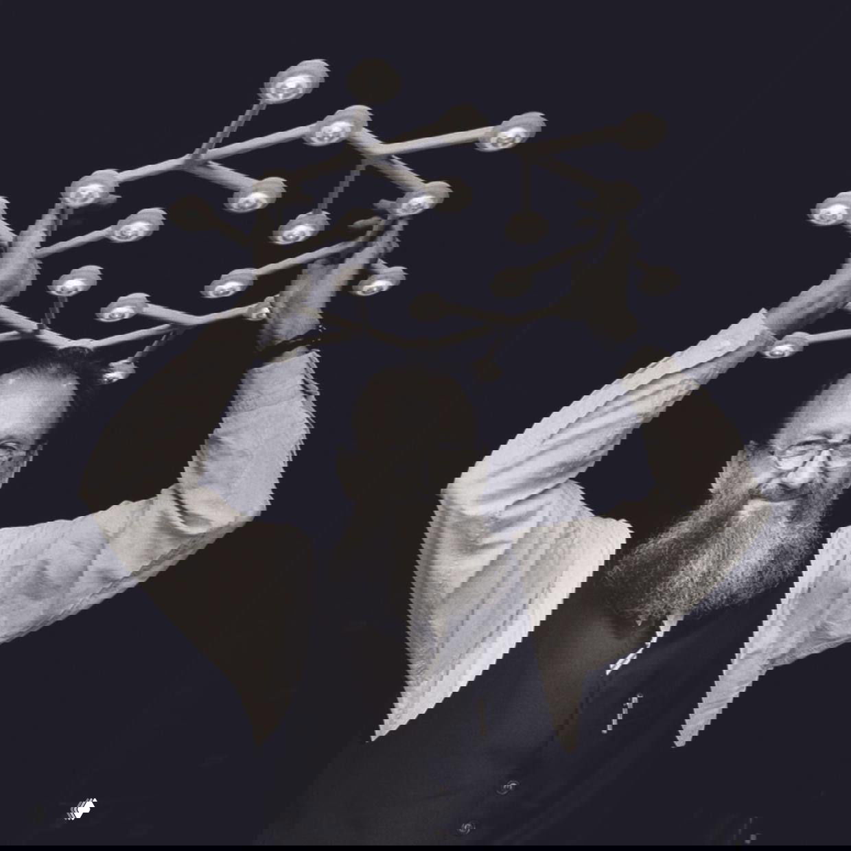 Чёрно-белый портрет улыбчивого дизайнера с бородой, держащего светильник над головой на тёмном фоне; акцент на форме светильника