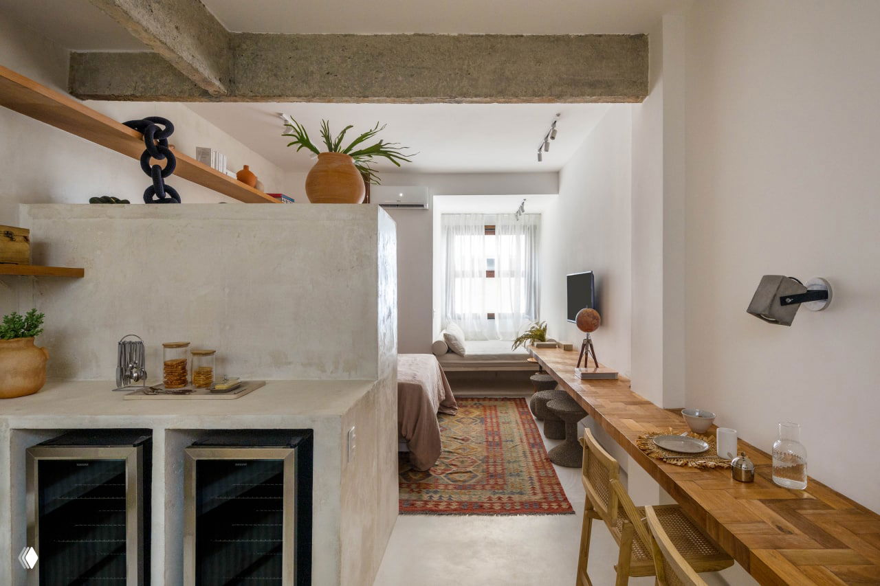 Широкий план компактной квартиры: объединённая кухня и спальня, бетонные элементы и деревянные поверхности, тёплый ковёр у окна создаёт уют.