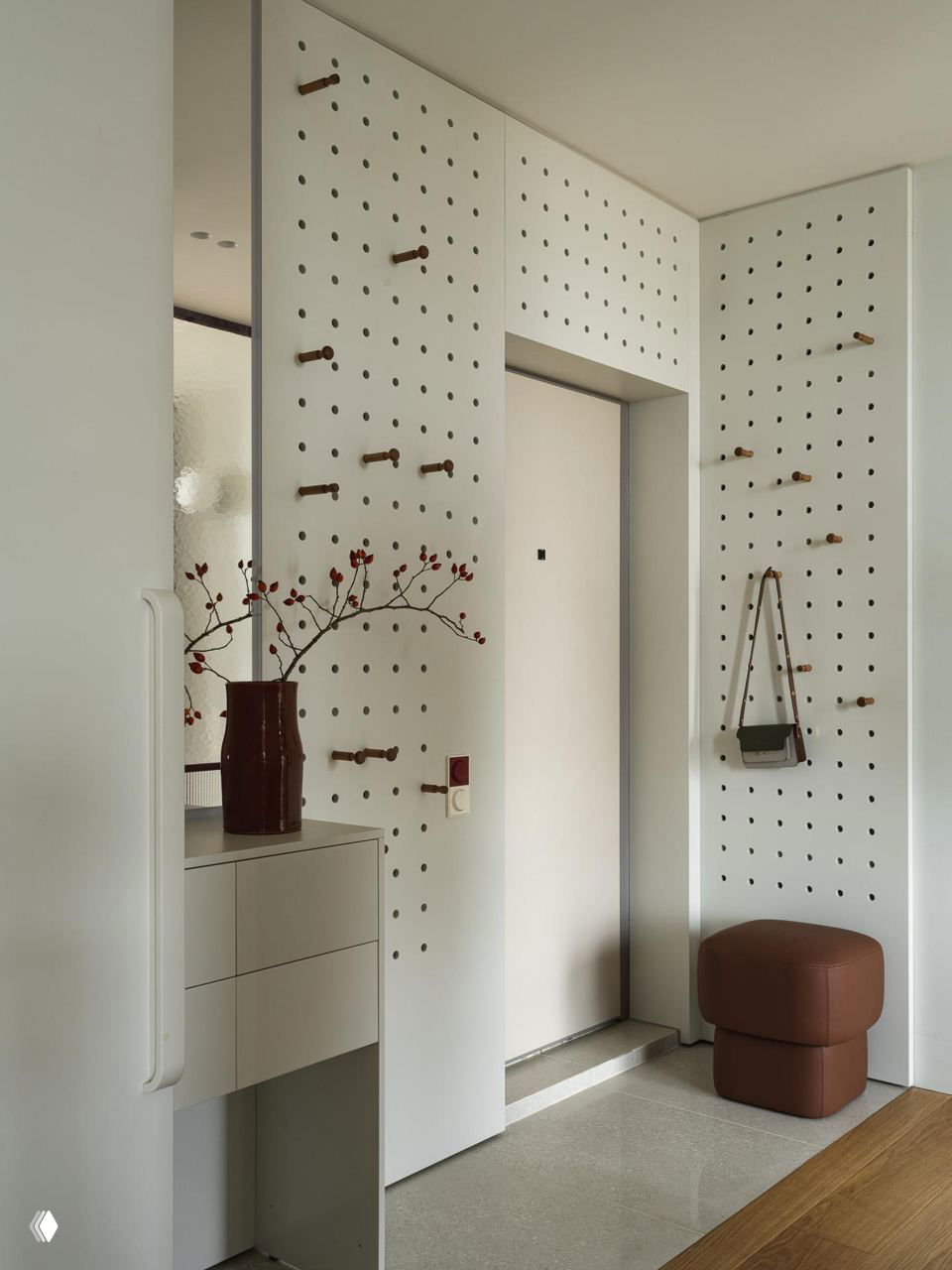Прихожая с перфорированными белыми панелями для кронштейнов, небольшим кожаным пуфом и вазой с веткой — система хранения на небольшой площади