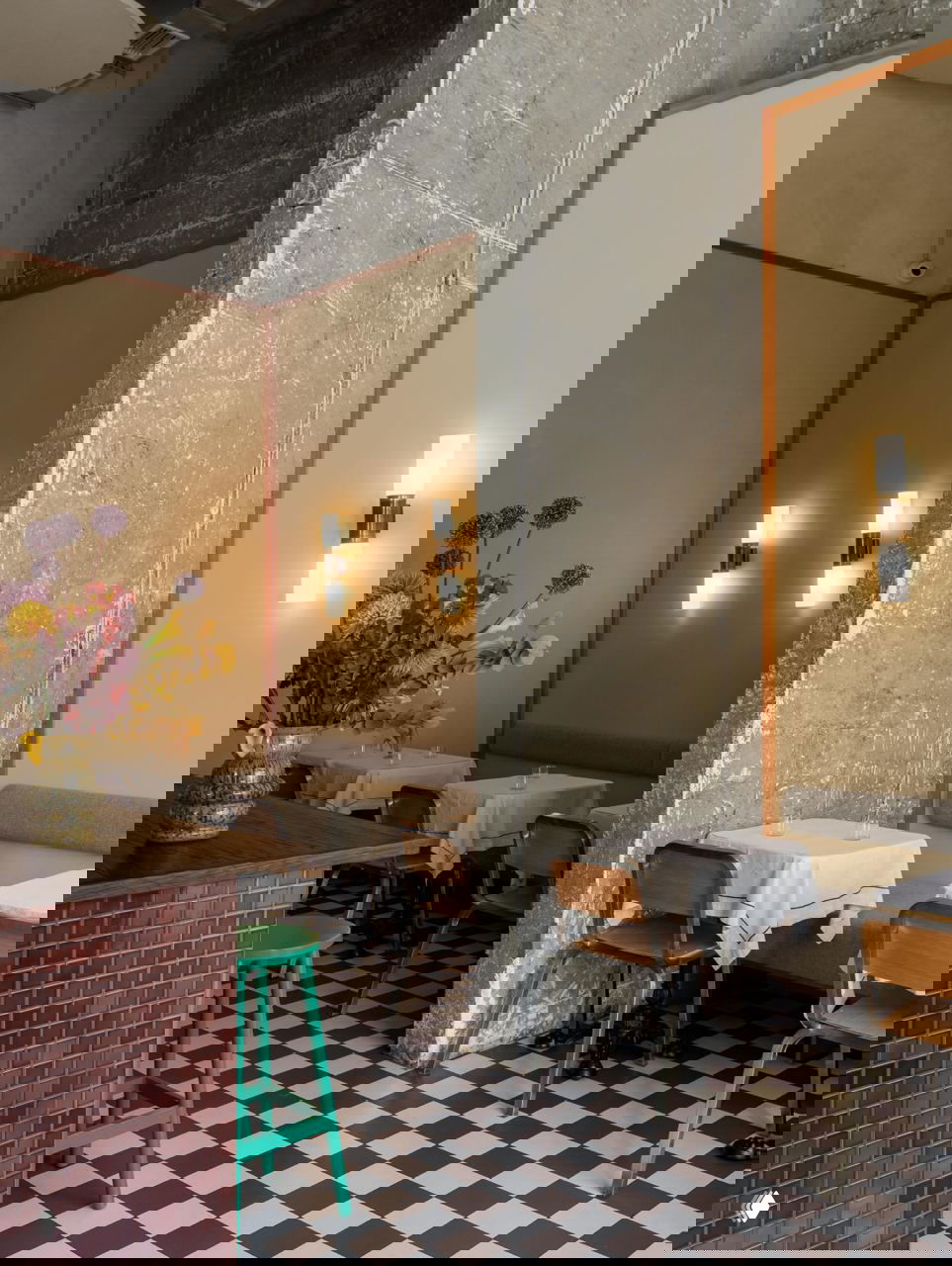 Узкая часть зала с бетонной колонной, настенными бра, мягкой скамьёй и небольшими столиками, плитка в шахматную клетку на полу.