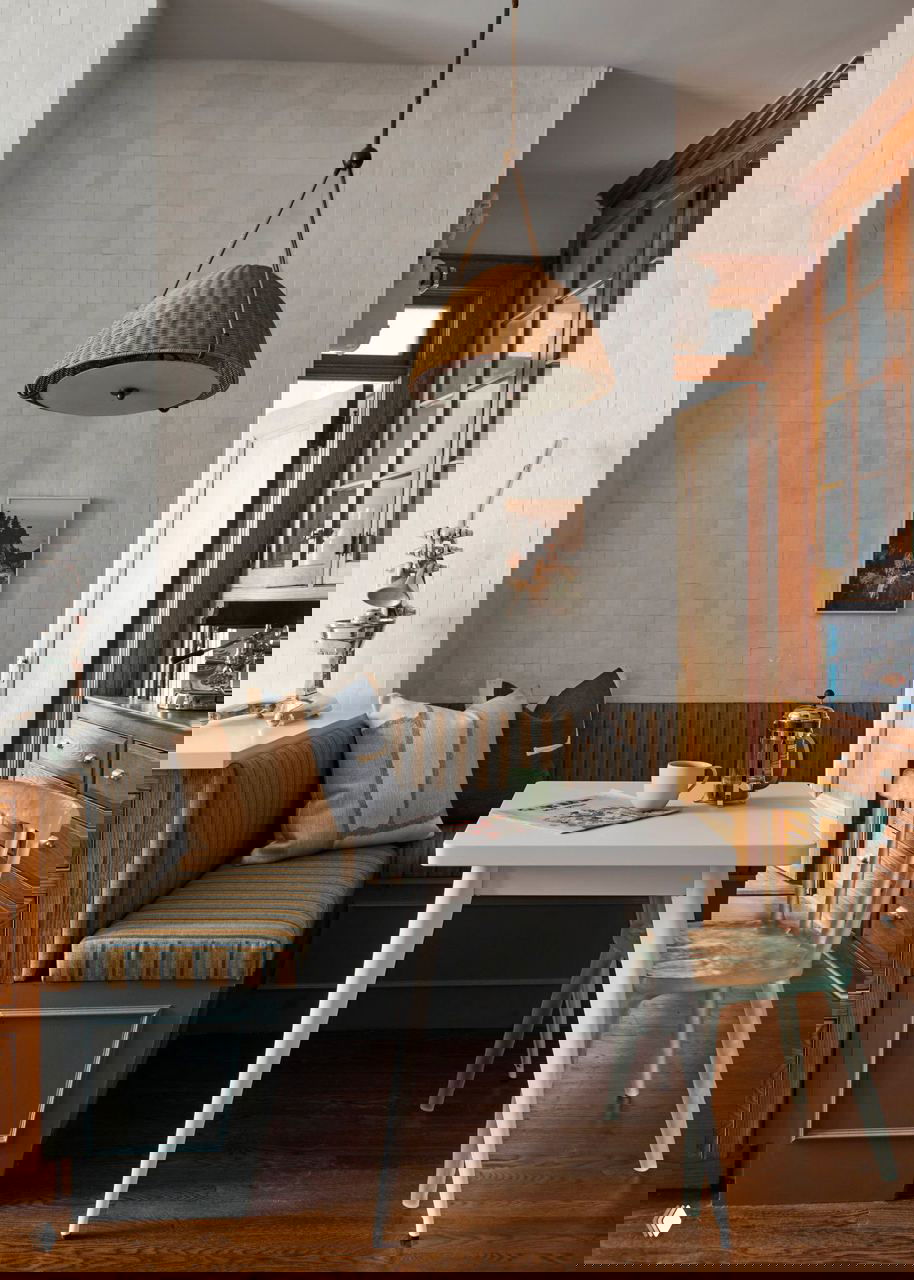 Небольшая обеденная зона со встроенным диваном и плетёным подвесным абажуром; уютный уголок рядом с деревянными шкафами.