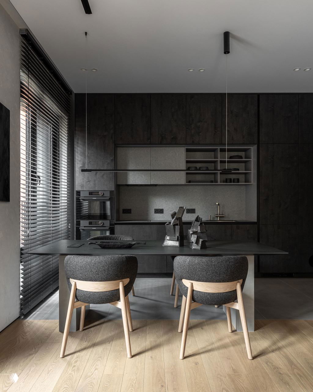Обеденная зона и рабочий стол перед тёмной кухней: деревянные стулья, низкая подвесная лампа и строгая монохромная отделка.