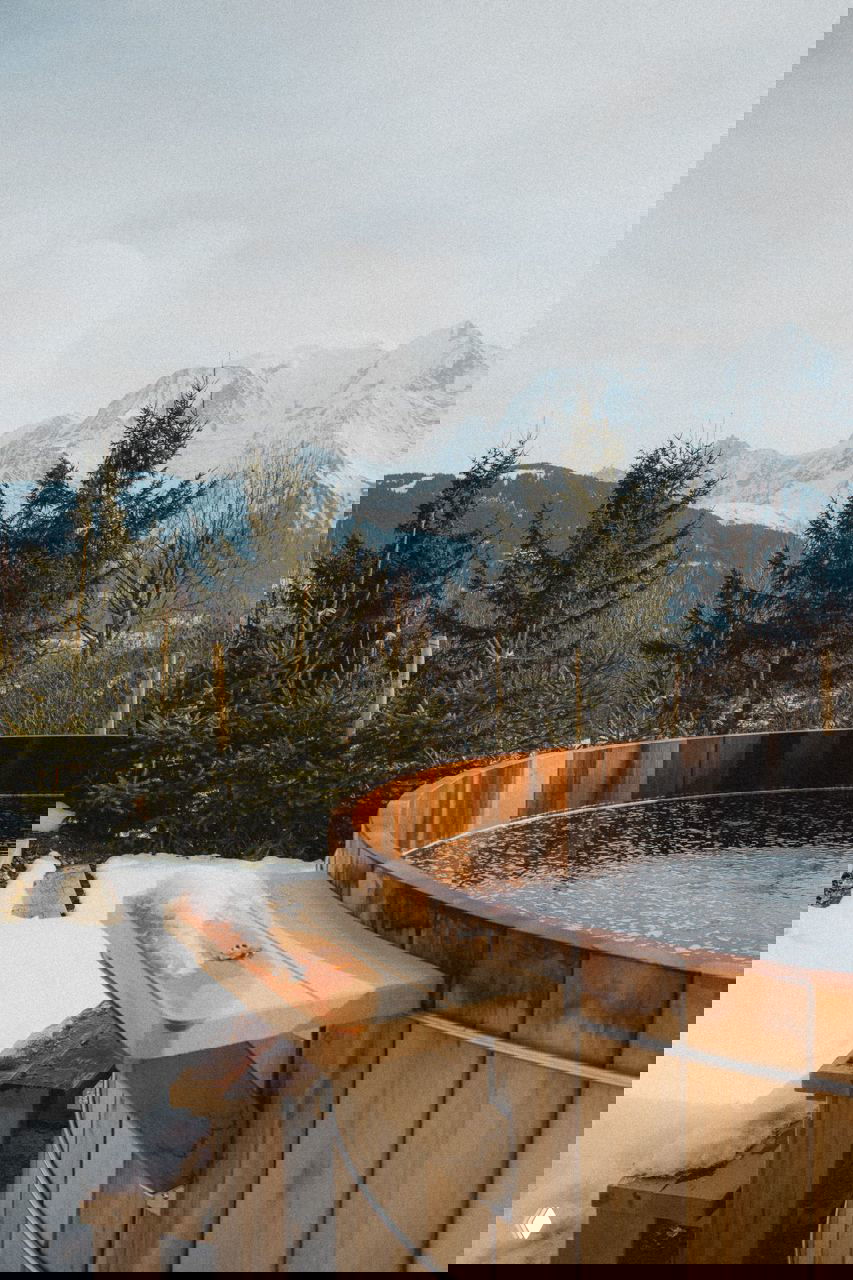 Купель или деревянная бочка для купания на снежном участке рядом с елями, вдалеке виднеются заснеженные вершины Монблана.