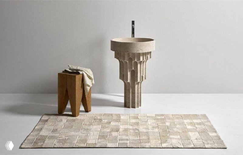 Раковина Carsico в интерьере: круглая верхняя чаша и снизу ритмичная решётка отверстий; рядом деревянный табурет и коврик, показана масштабность изделия.