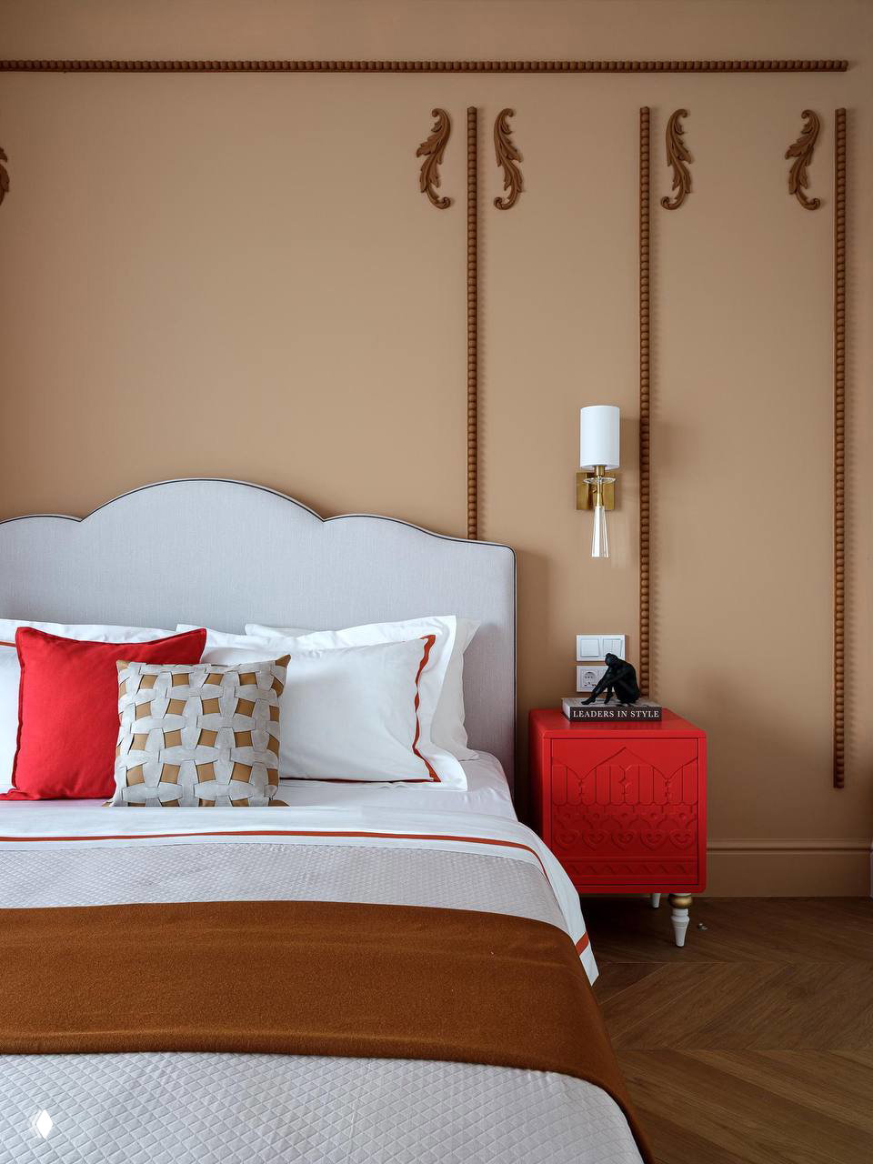 Спальня в теплых бежевых тонах: изголовье кровати светлое, текстиль с акцентом красного и прикроватный комод насыщенного алого цвета.