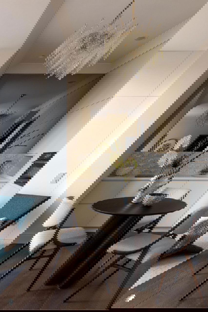 Столовая и мини‑кухня в светлой палитре: круглый стол, изящная люстра и зеркало за столовой зоной — уютная планировка проекта Ольги Рябовой.