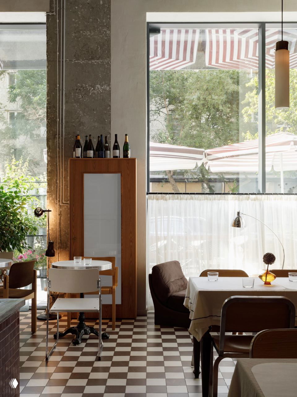 Вид на большое окно с тентовыми навесами снаружи, в интерьере — столики у окна, деревянные и мягкие кресла, дневной свет наполняет зал.