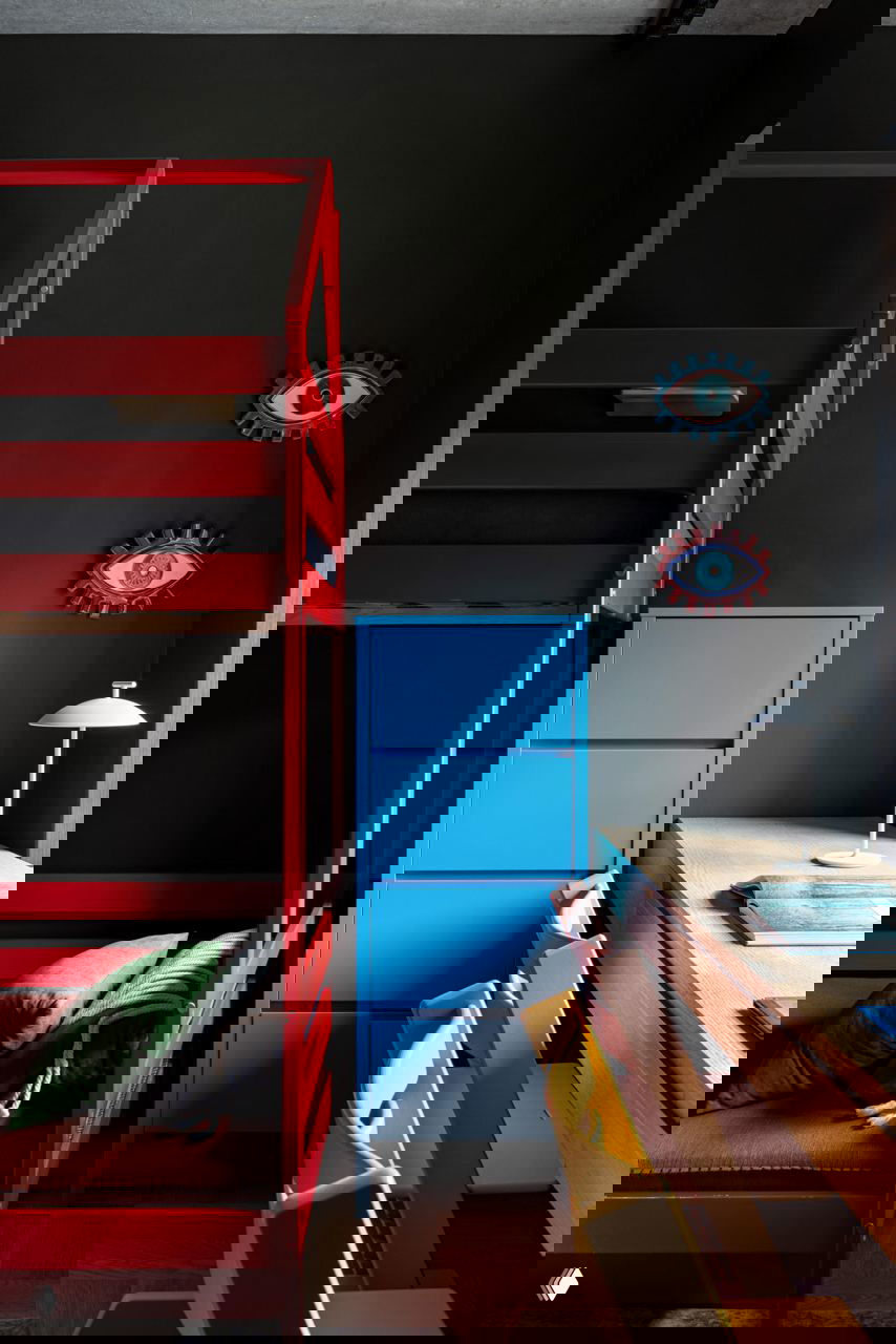 Детская комната с двухъярусной кроватью красного цвета, голубым комодом и рабочей зоной с яркими стульями на фоне темных стен.