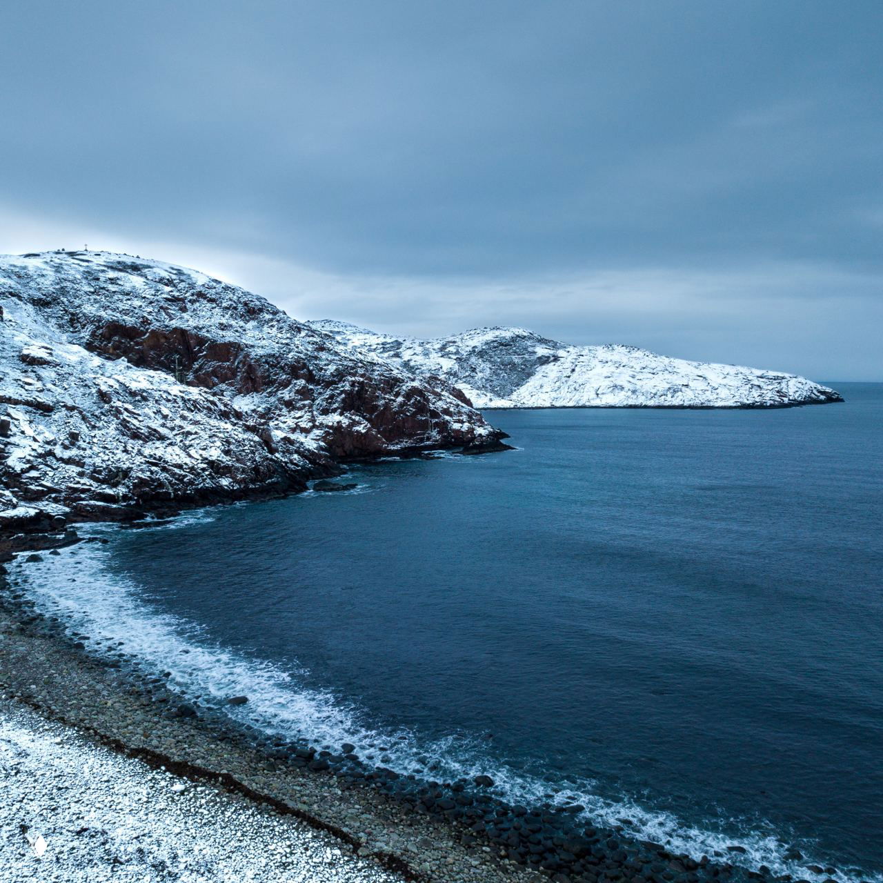 Каменный и снежный берег Баренцева моря у Териберки: суровые скалы, холодная морская гладь и прибрежная галька под низким зимним небом.