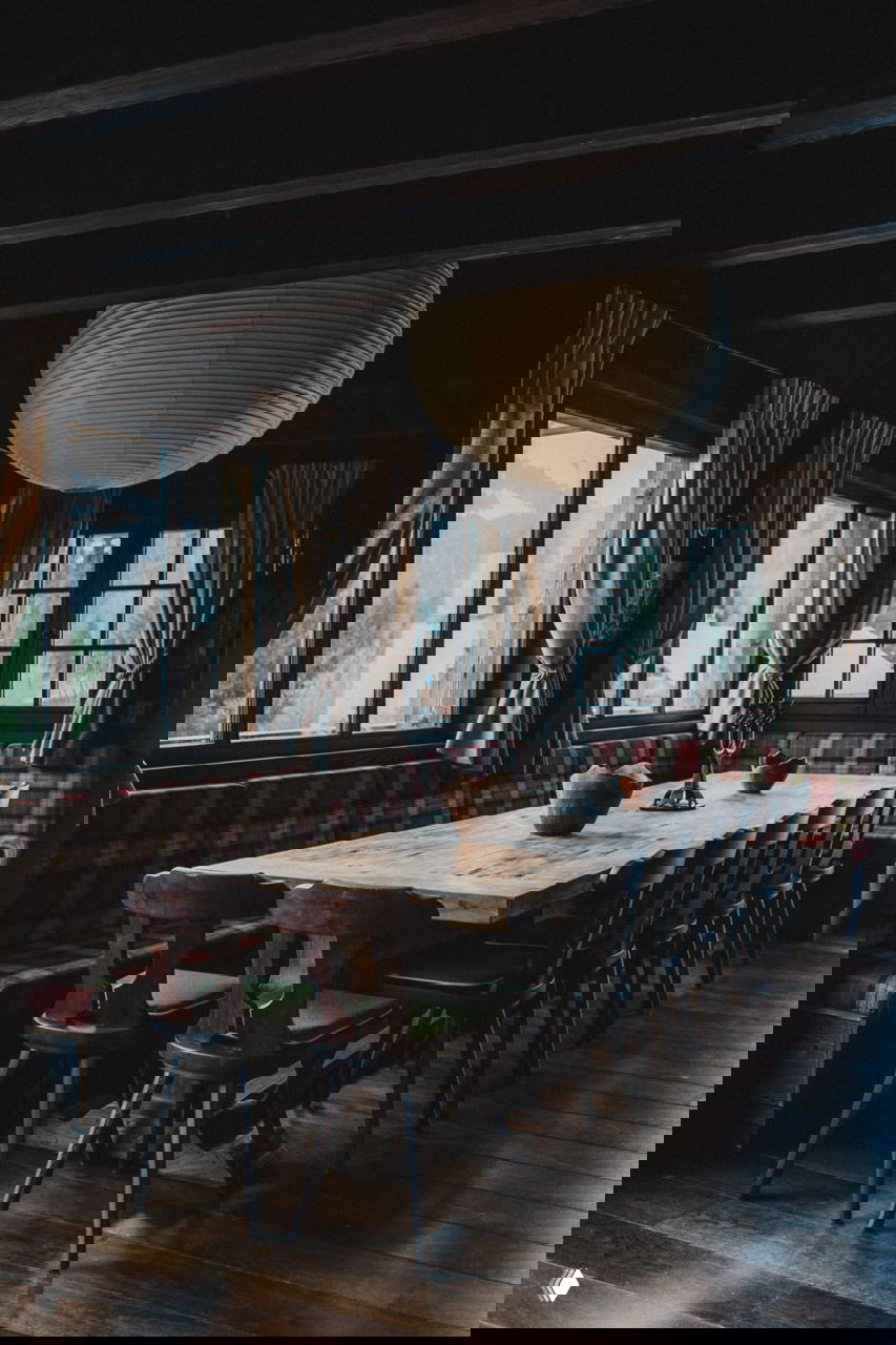 Столовая шале с длинным массивным деревянным столом, скамьёй в клетчатой обивке и высокими окнами, наполняющими пространство естественным светом.