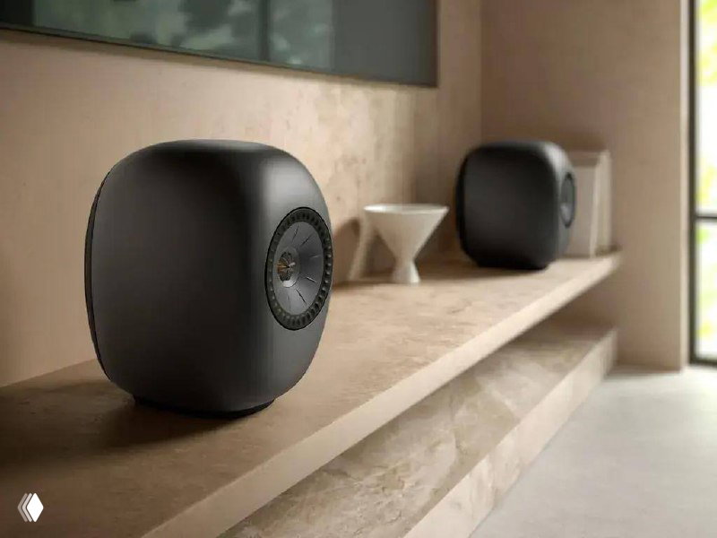 Матовая чёрная колонка KEF округлой формы на каменной полке в интерьере, пара колонок на заднем плане и естественное освещение